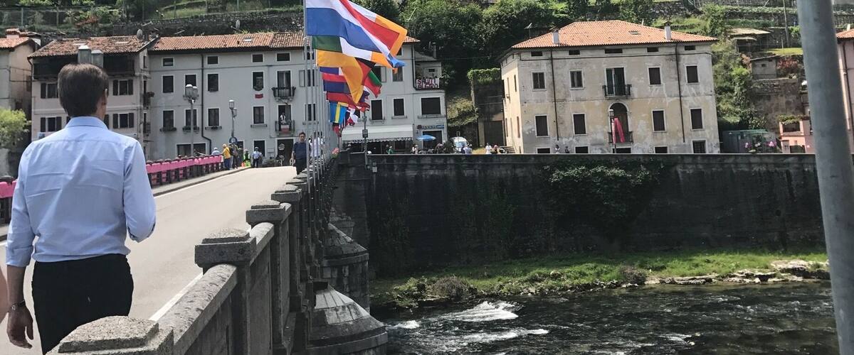 A man walking on a bridge with a colorful flag, a river below, and buildings with red-tiled roofs in a mountainous area.