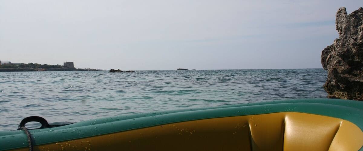 A boat on calm sea waters with a rocky outcrop on the right and a distant shoreline with buildings.