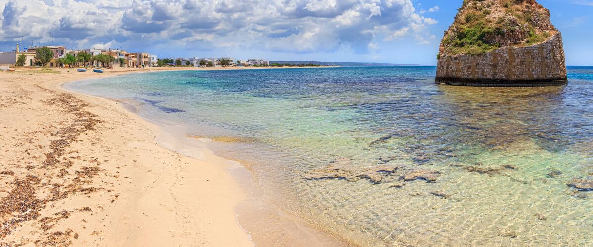 A sandy beach with clear water, a rocky outcrop, and buildings in the background.