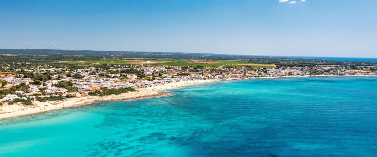 An aerial view of a coastal town with a beach, clear turquoise waters, and a distant shoreline.