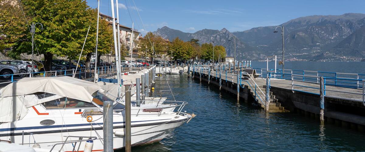 A marina with boats docked, a pier extending into the water, and mountains in the background.