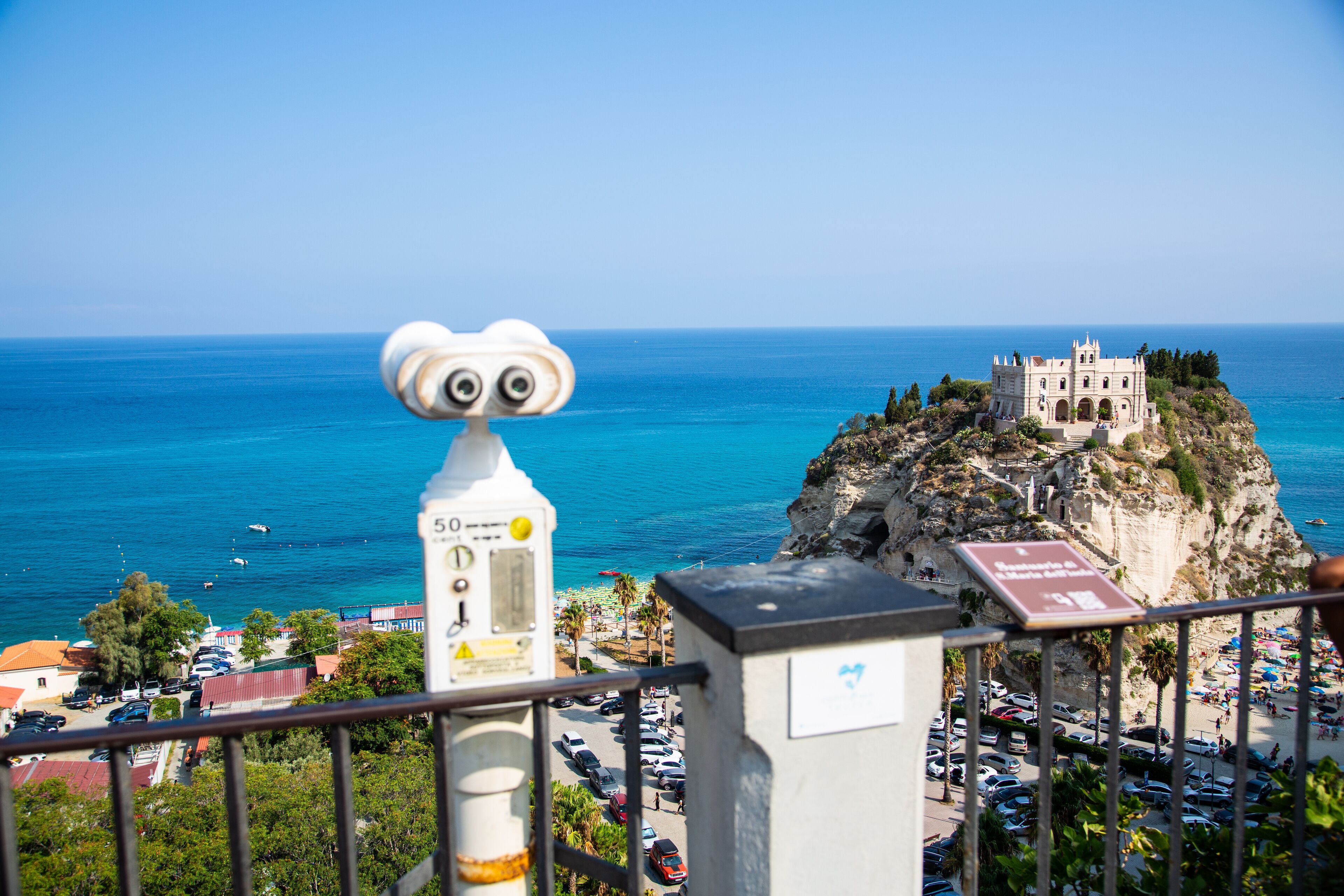 A coastal town with a historic building on a cliff, a lighthouse, and a beach with parked cars.