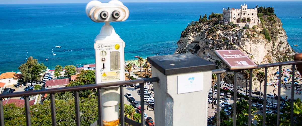 A coastal town with a historic building on a cliff, a lighthouse, and a beach with parked cars.