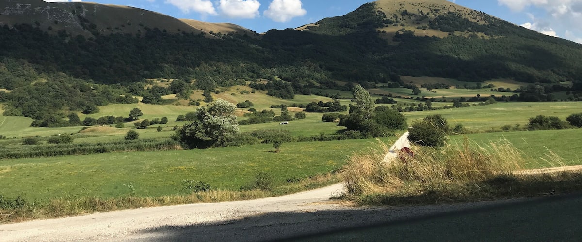 A rural landscape with rolling hills, a dirt road, and a clear blue sky with scattered clouds.