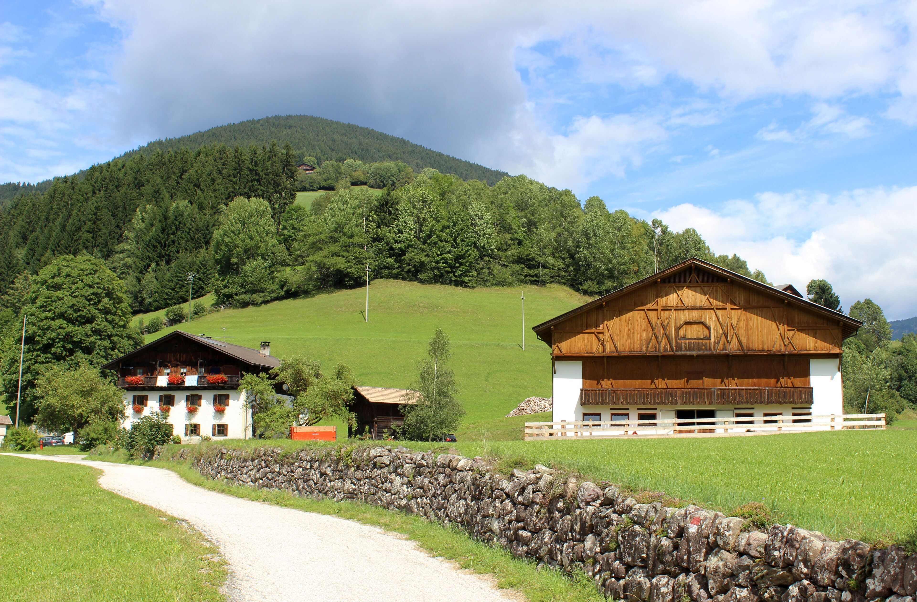 A rural landscape with a stone wall, a white building with red flower boxes, and a wooden barn.