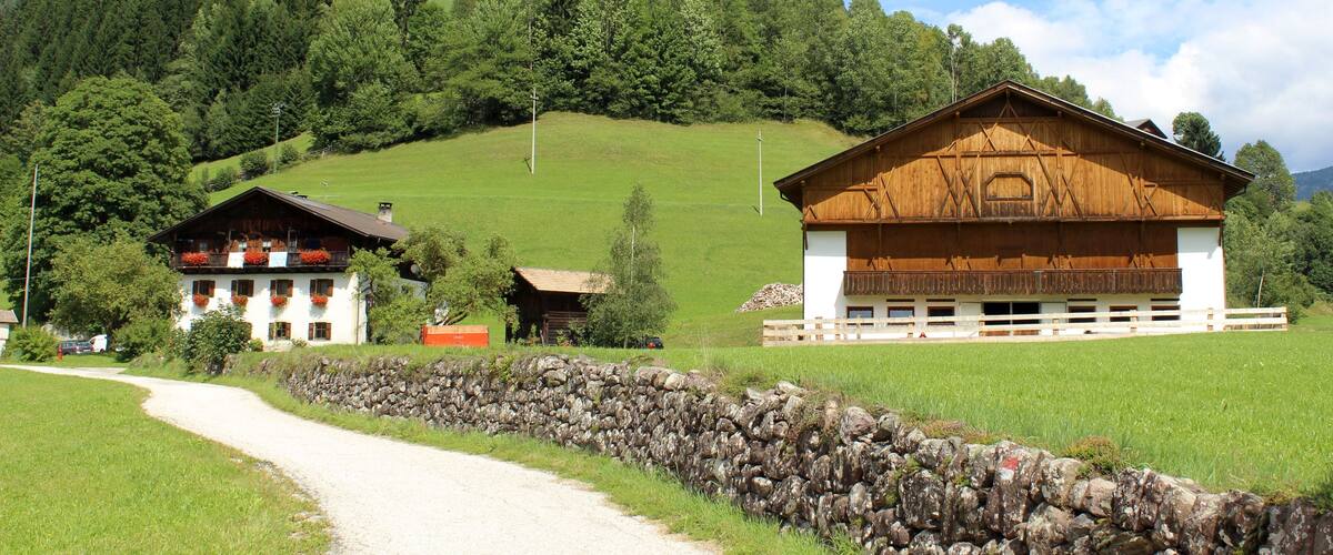 A rural landscape with a stone wall, a white building with red flower boxes, and a wooden barn.