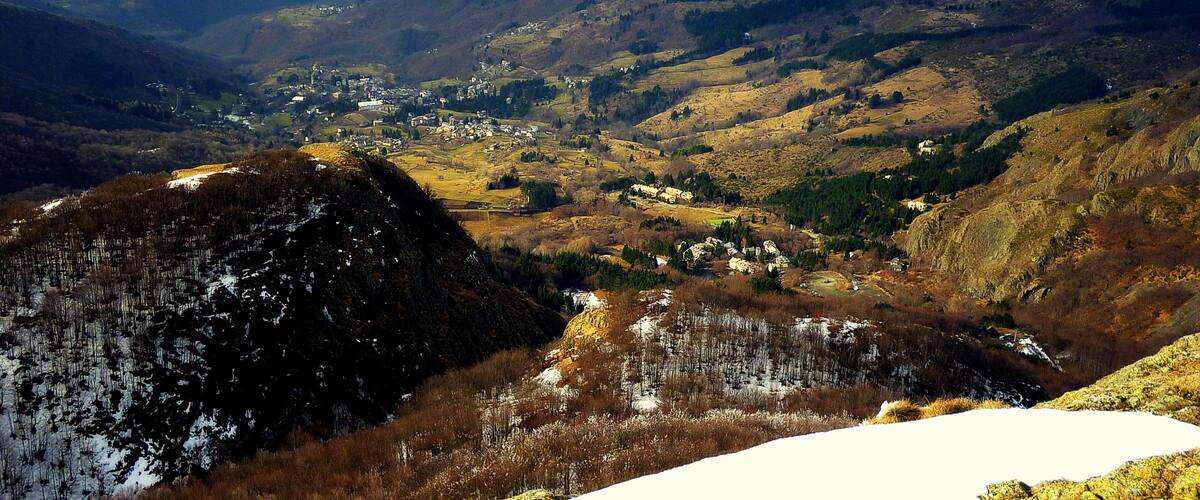 A mountainous landscape with a village nestled in the valley, snow patches, and distant mountain ranges.