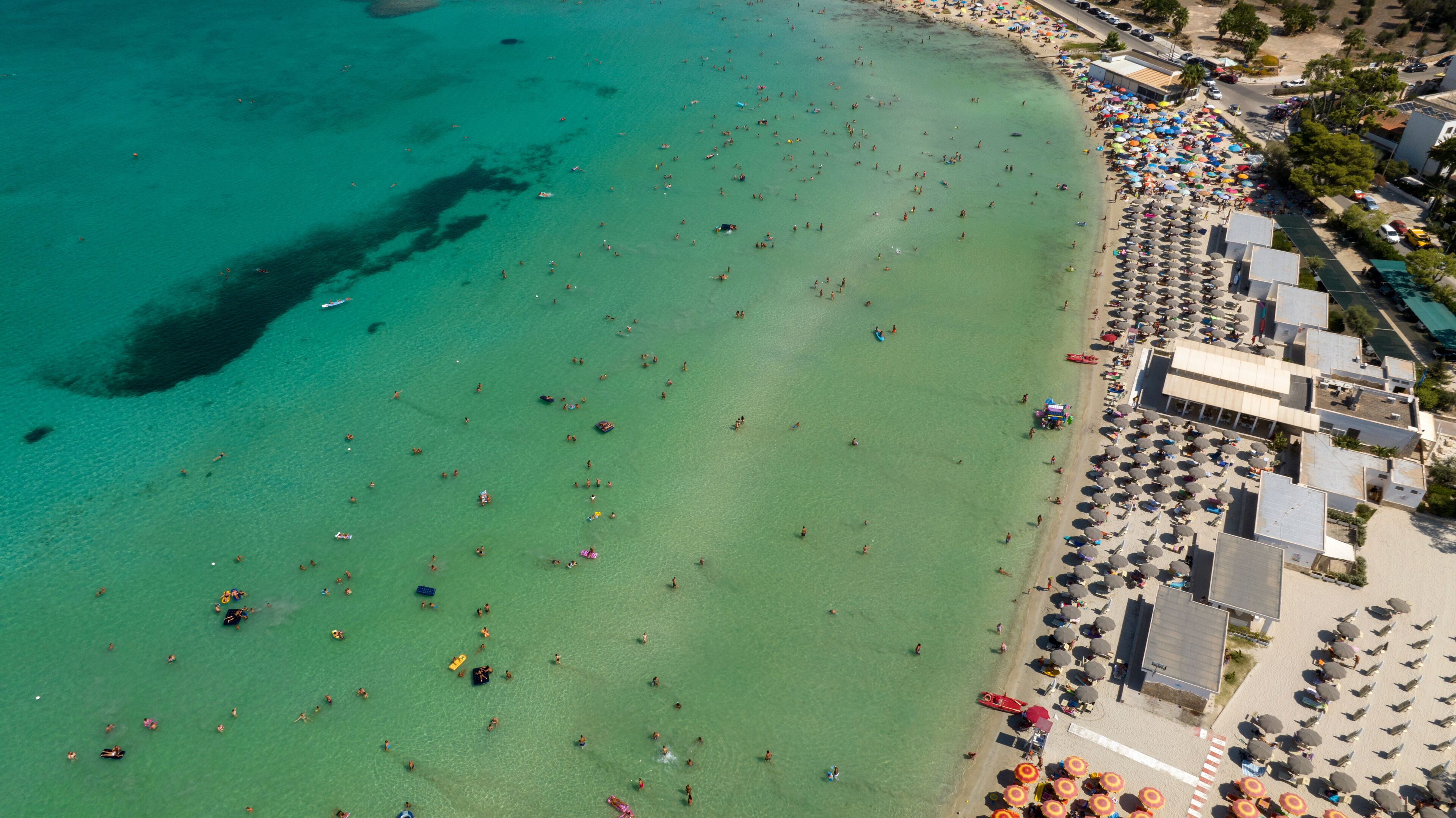An aerial view of a beach with clear turquoise waters, numerous umbrellas, and people enjoying the seaside.