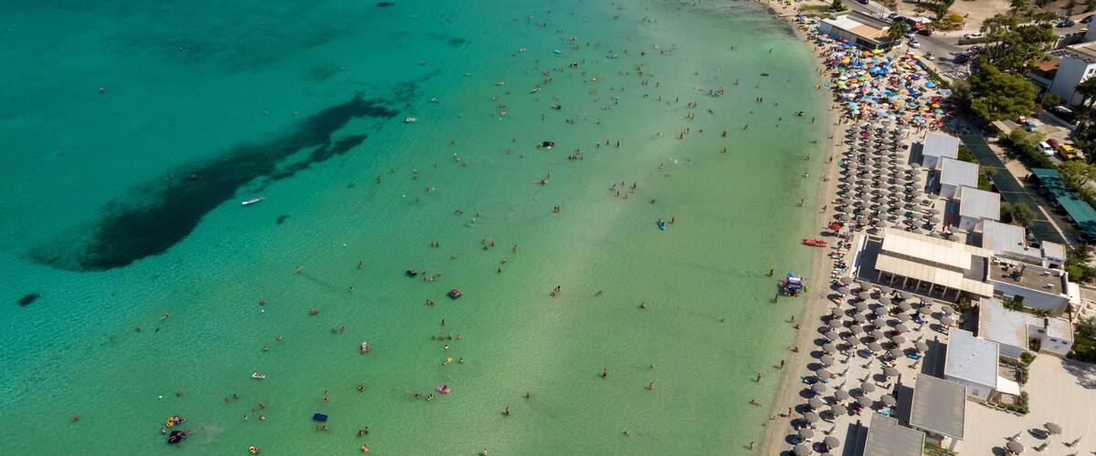 An aerial view of a beach with clear turquoise waters, numerous umbrellas, and people enjoying the seaside.