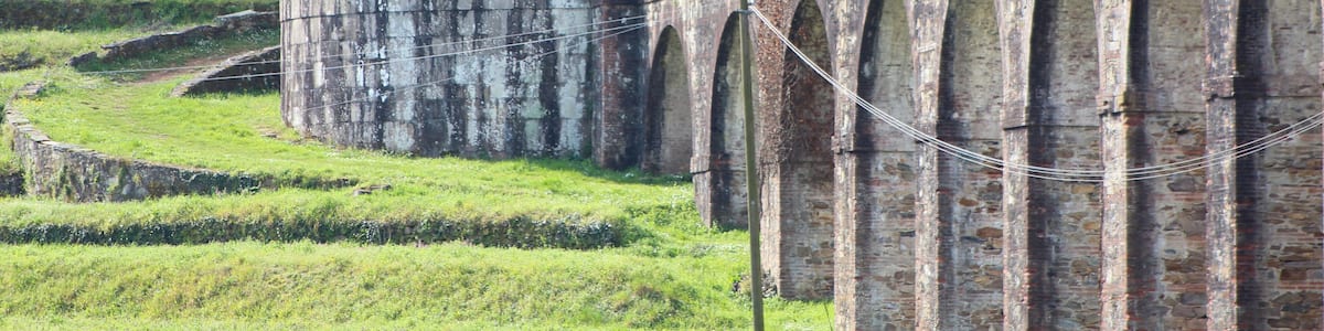 Un pont en pierre, doté d’arches et surmonté d’une structure en forme de dôme avec une petite ouverture, le tout entouré de verdure et d’arbres.