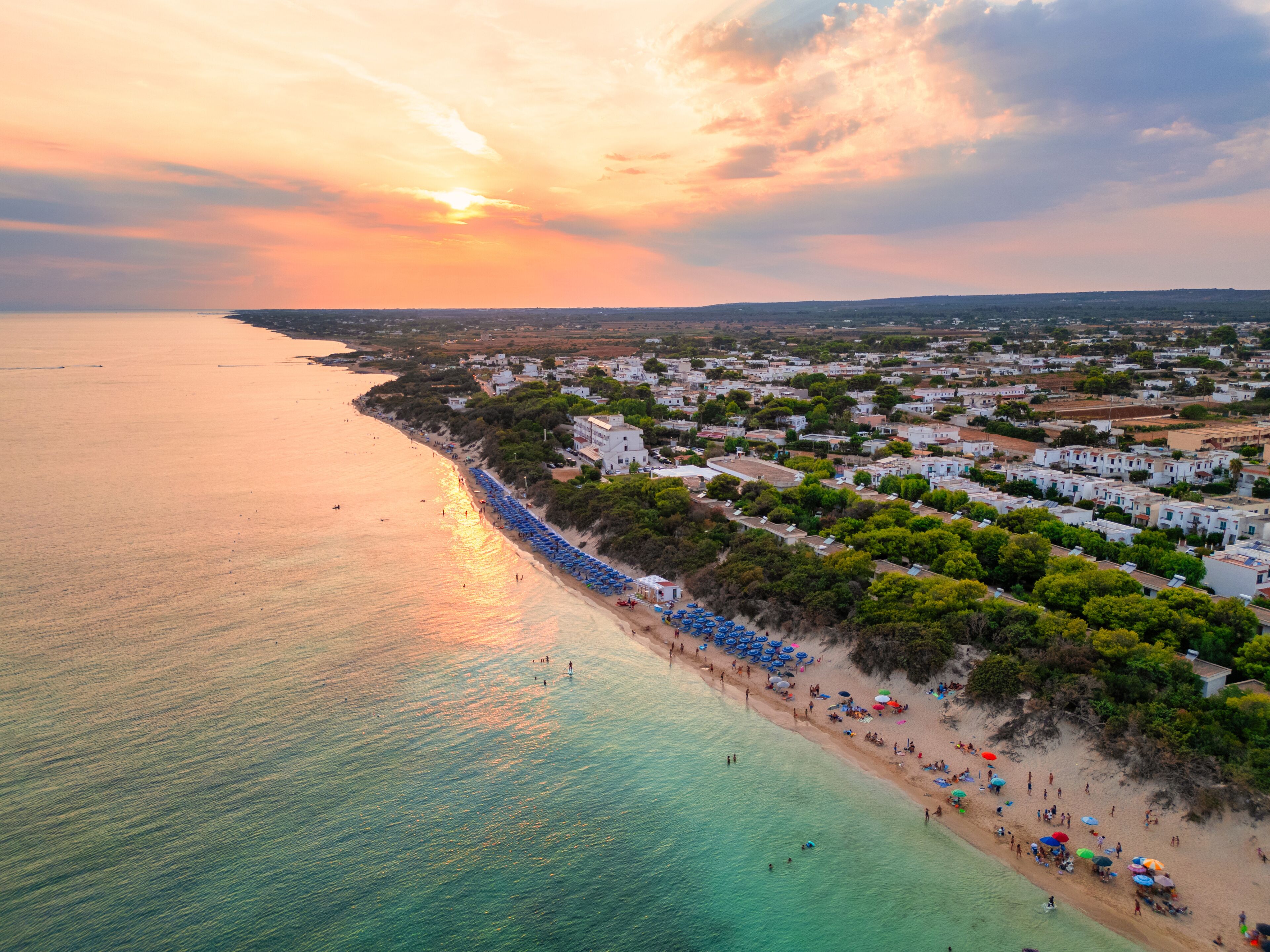 A coastal town with a beach lined with umbrellas and people, surrounded by white buildings and greenery.