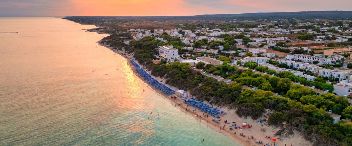 A coastal town with a beach lined with umbrellas and people, surrounded by white buildings and greenery.