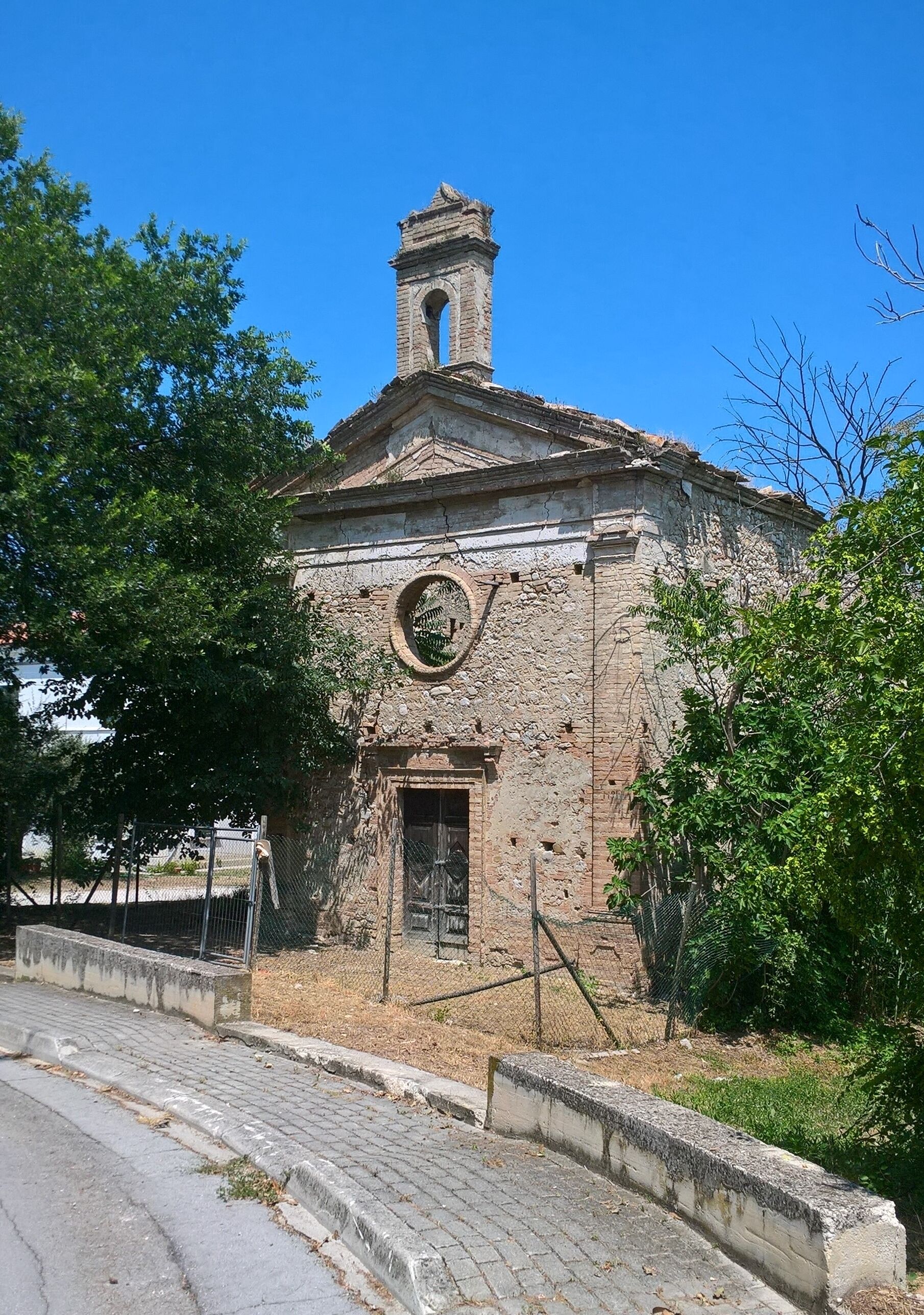 An old stone church with a bell tower, surrounded by trees and a paved road.