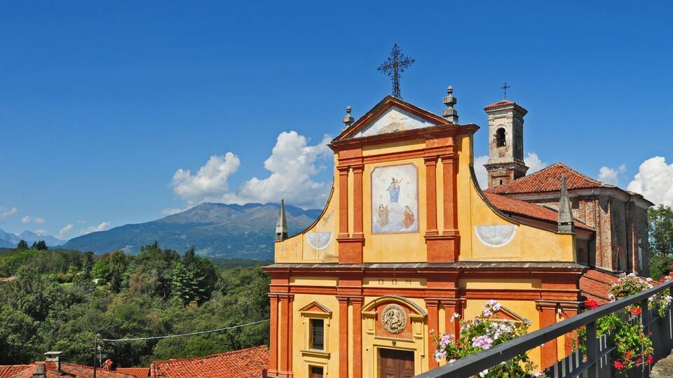 A church with a bell tower and a cross on top, surrounded by mountains and greenery.