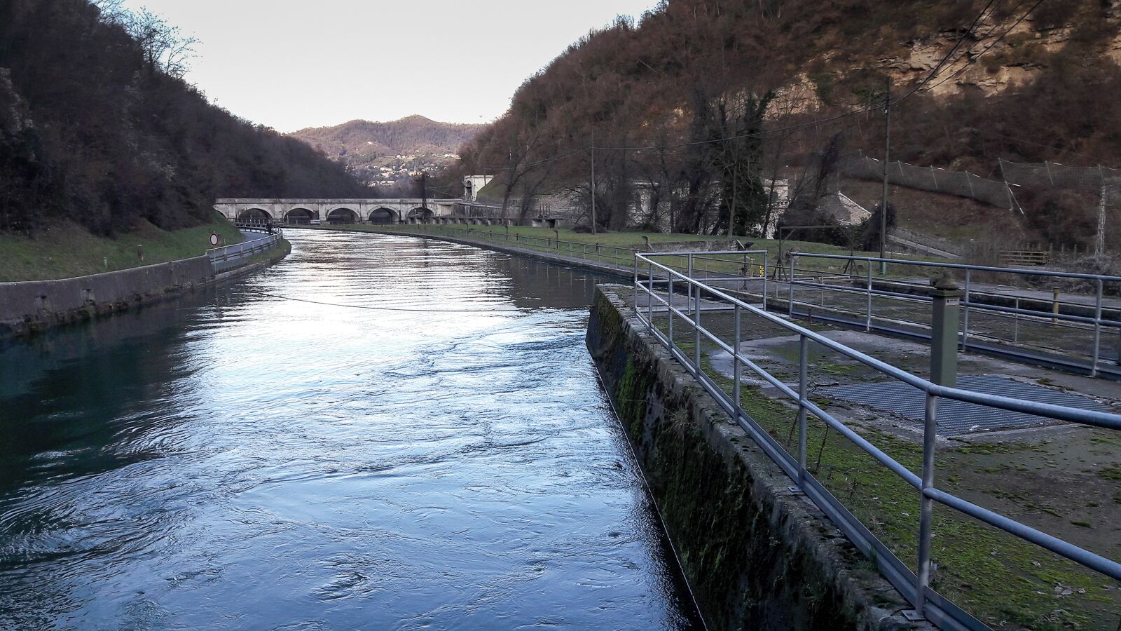 A canal with a bridge in the background, surrounded by hills and trees.
