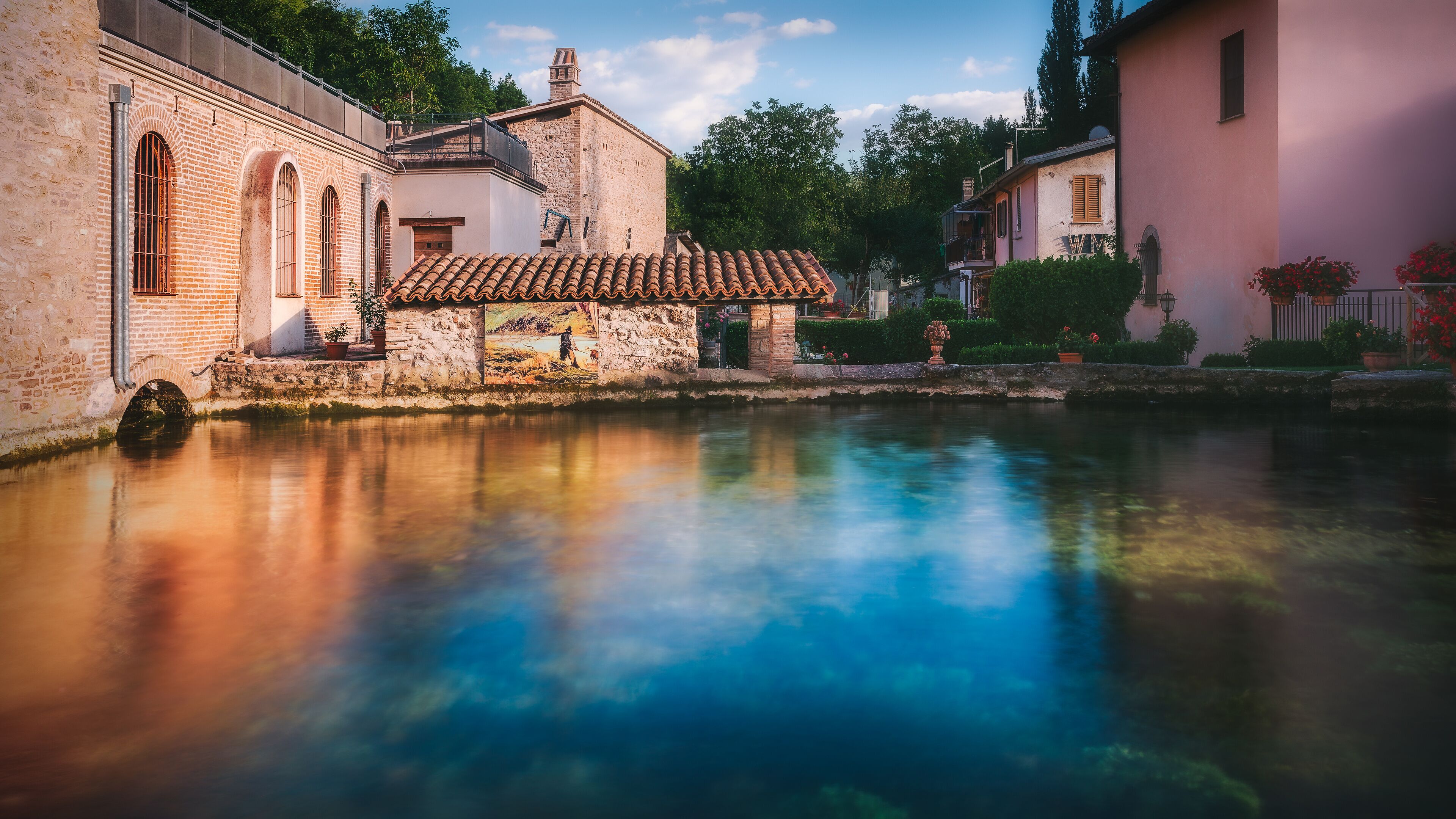 A historic building with a tiled roof and a stone wall, reflecting in a calm water body.
