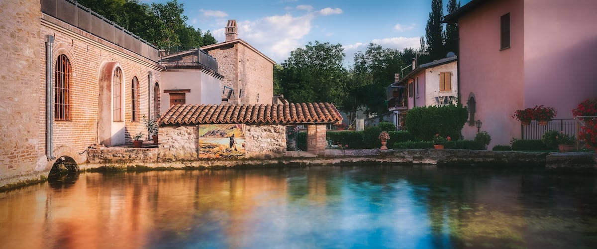 A historic building with a tiled roof and a stone wall, reflecting in a calm water body.