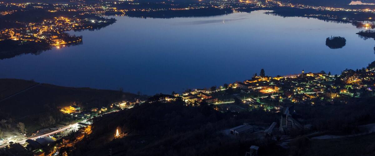 A panoramic view of a city at dusk with a large body of water in the center, surrounded by illuminated buildings and a hilly landscape.