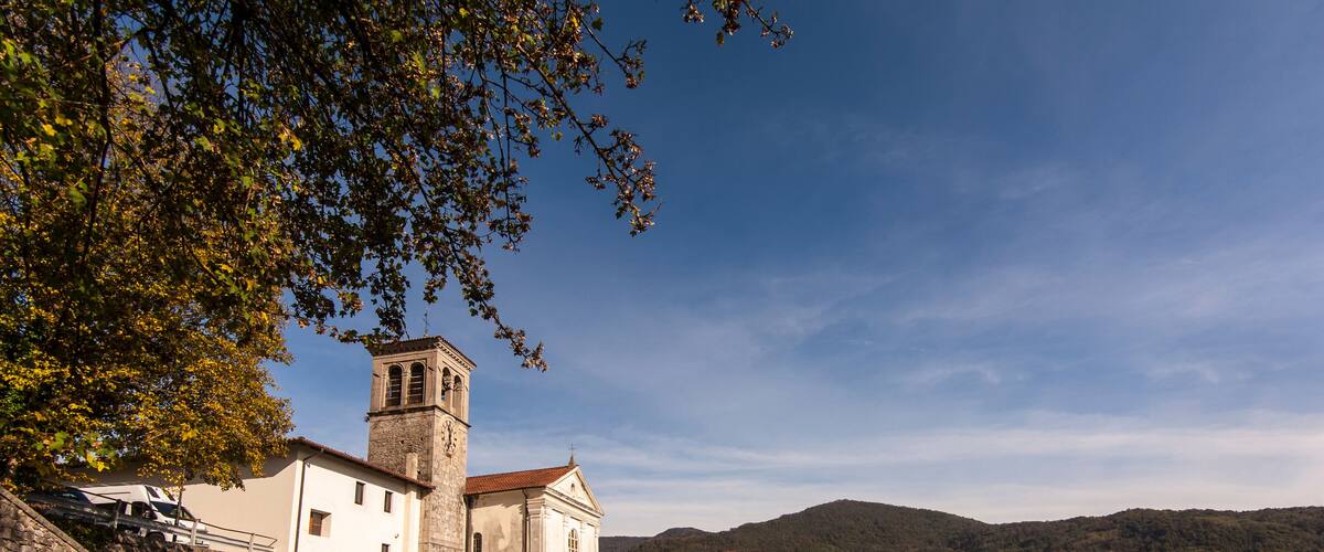 A historic stone church with a bell tower, surrounded by traditional buildings and a mountainous backdrop.