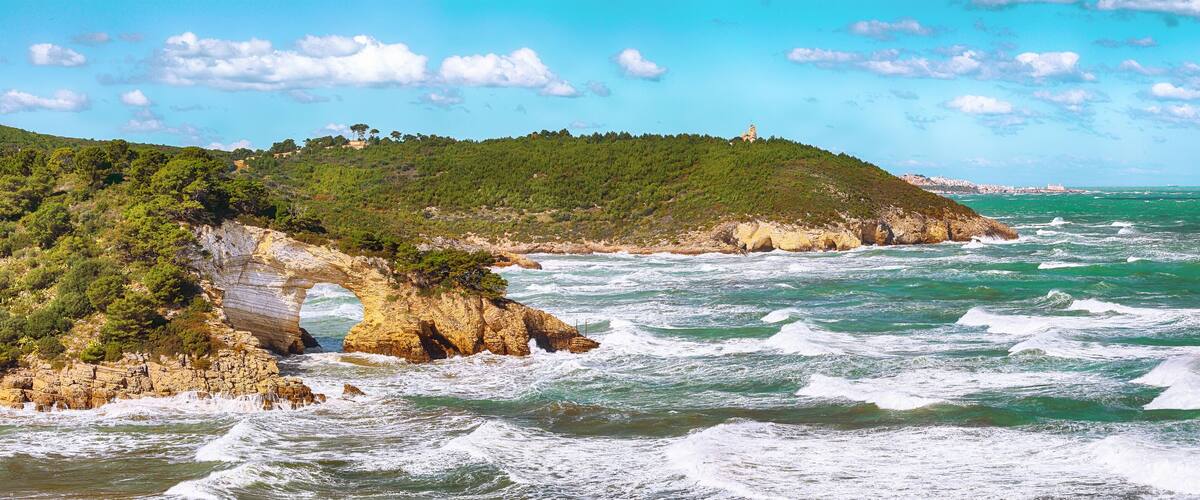 A coastal landscape with rocky cliffs, a clear blue sky, and waves crashing against the shore.