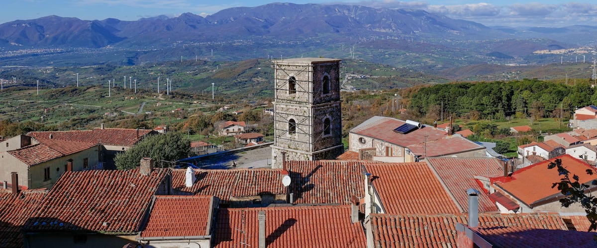 A town with red-tiled roofs, a prominent tower, and mountains in the background.