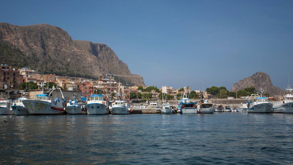 A harbor with several boats docked, a mountainous backdrop, and buildings along the waterfront.