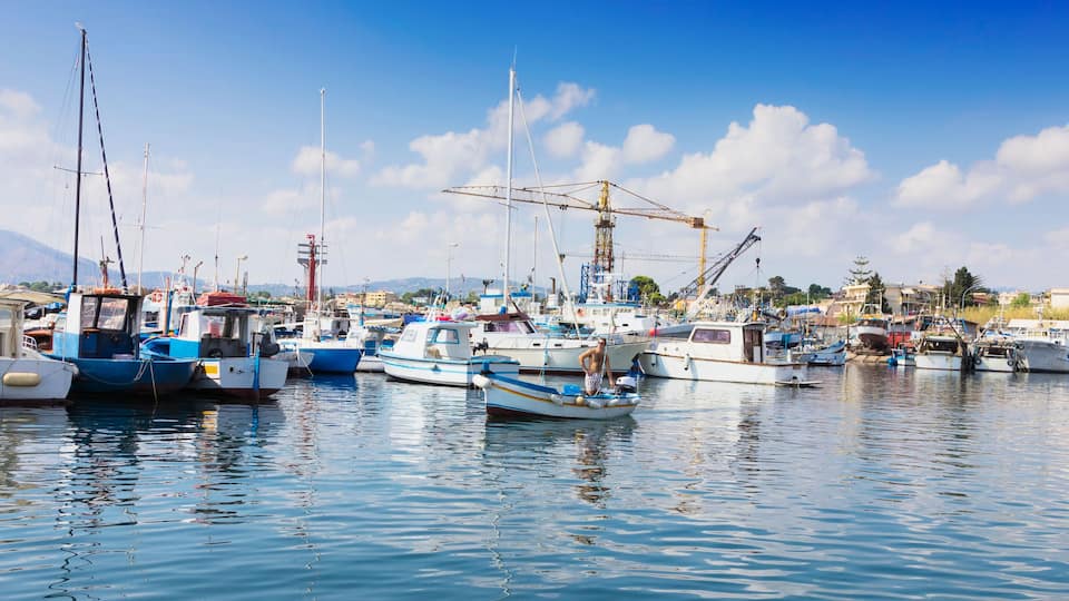 A marina with various boats docked, a crane, and mountains in the background.