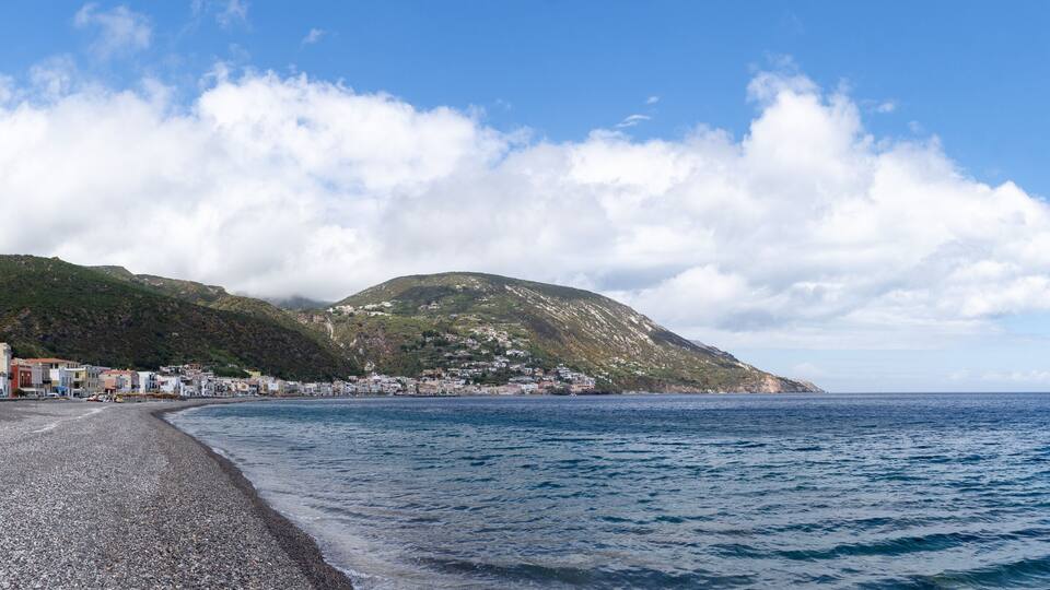 A pebble beach with buildings and a mountain in the background.