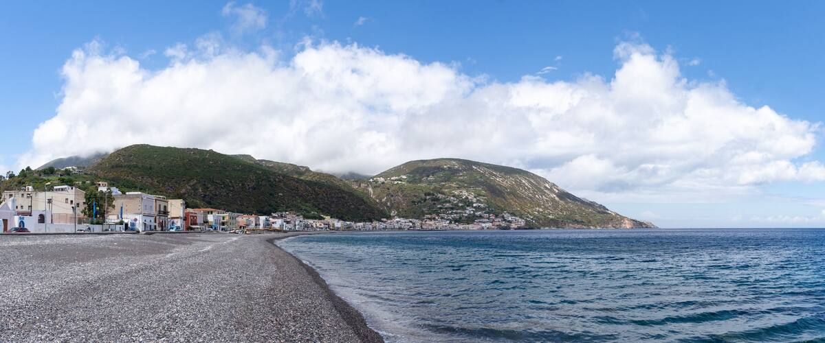 A pebble beach with buildings and a mountain in the background.