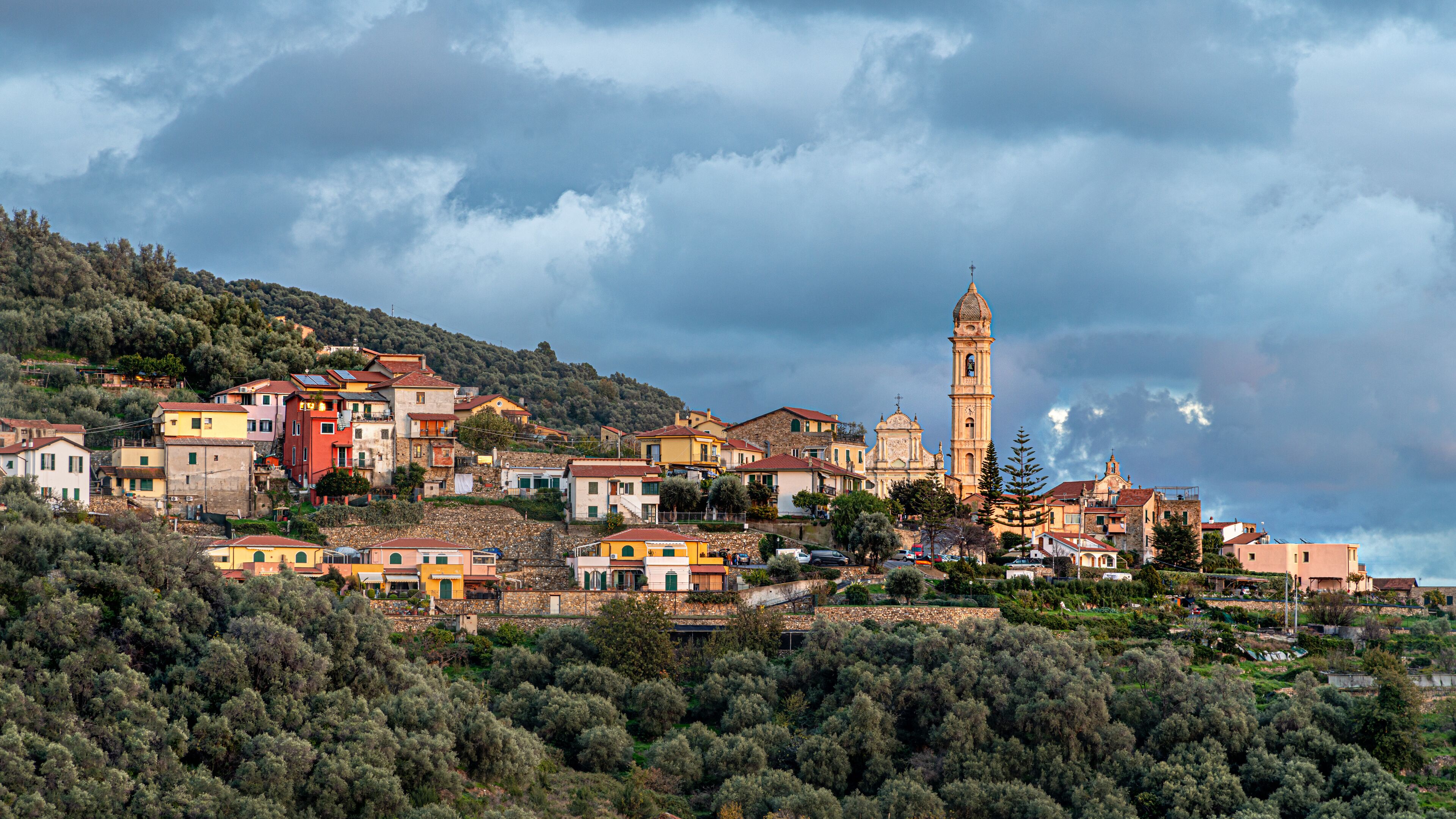 A coastal town with colorful buildings, a prominent church with a bell tower, and a backdrop of hills and trees.