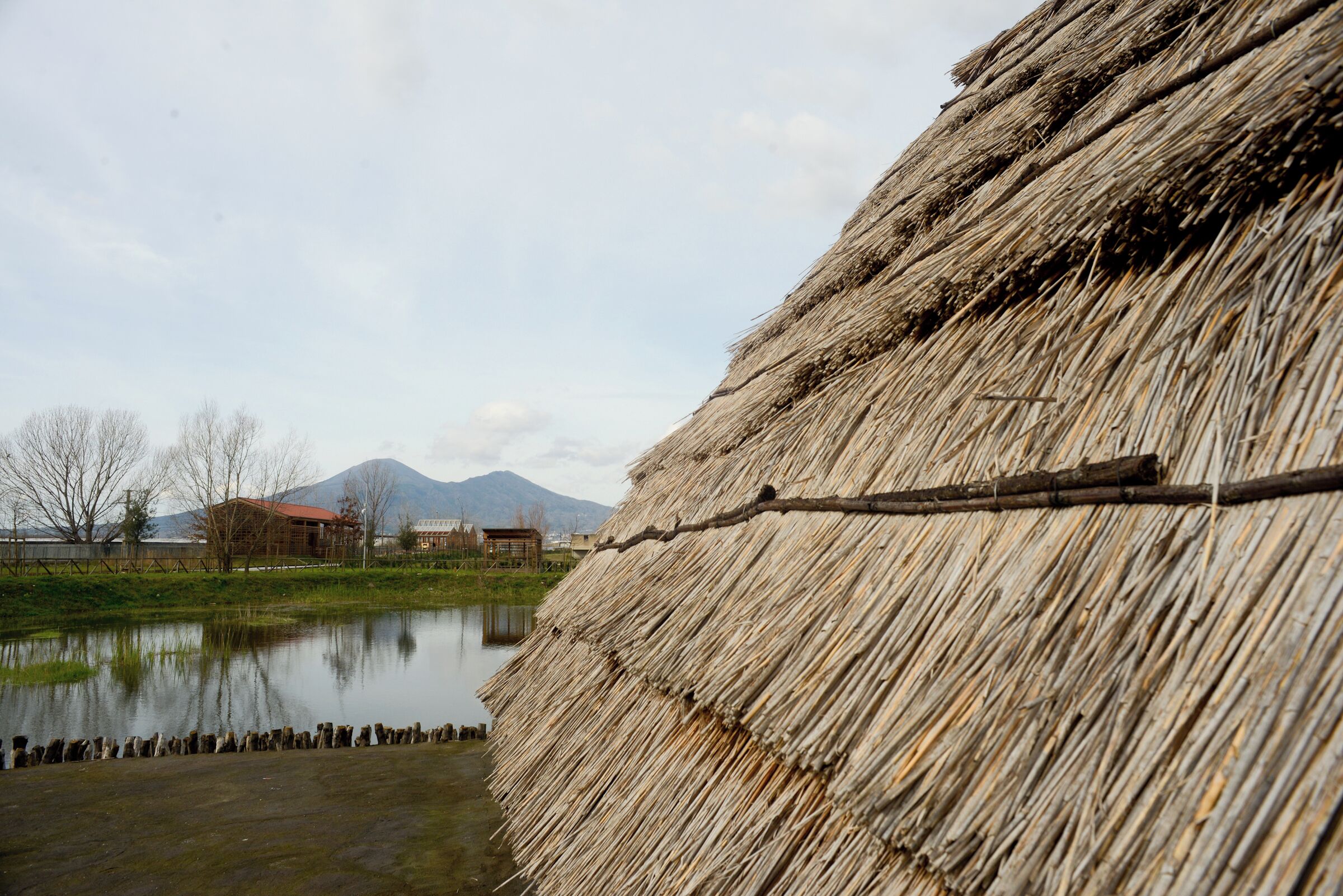 A thatched structure by a calm pond with mountains in the background.