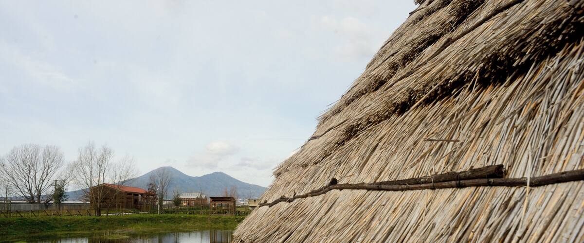 A thatched structure by a calm pond with mountains in the background.