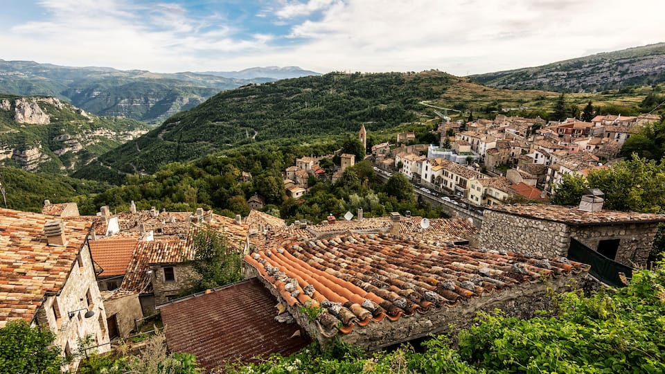 A historic town with terracotta-roofed buildings, a stone amphitheater, and lush green hills in the background.