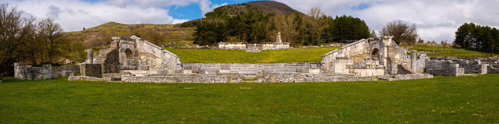 Ancient stone ruins with a grassy field in the foreground and a hill with trees in the background.