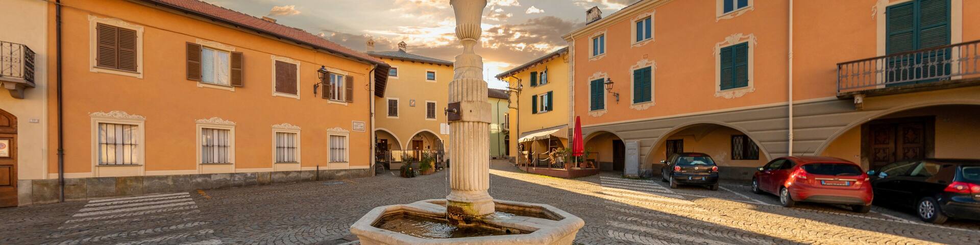 Peveragno, Cuneo, Italy - January 09; 2023: marble fountain with drinking trough on cobblestones in Piazza Santa Maria with ancient palaces decorati with arcade under cloudy sky at sunset