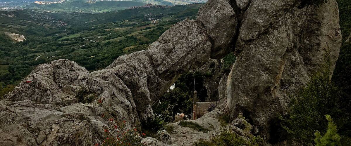 A rocky landscape with a prominent rock formation in the foreground and a view of a valley with scattered buildings in the distance under a cloudy sky.