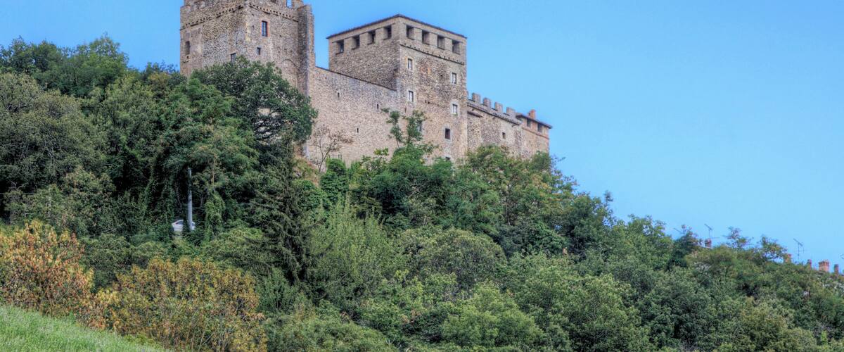 A historic stone castle with a tower and a smaller building, surrounded by dense green trees.