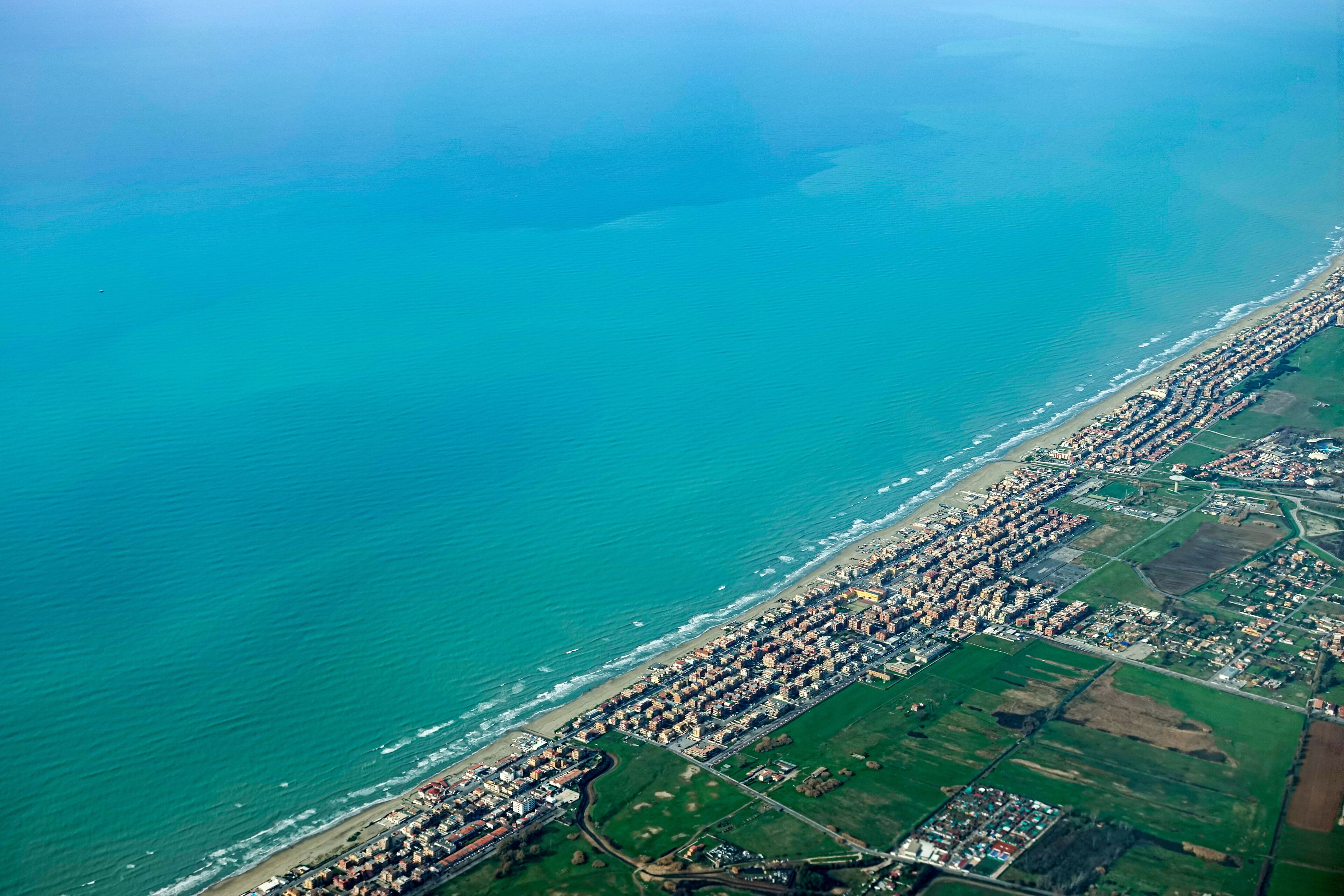 An aerial view of a coastal city with a long beachfront, dense residential area, and green fields.