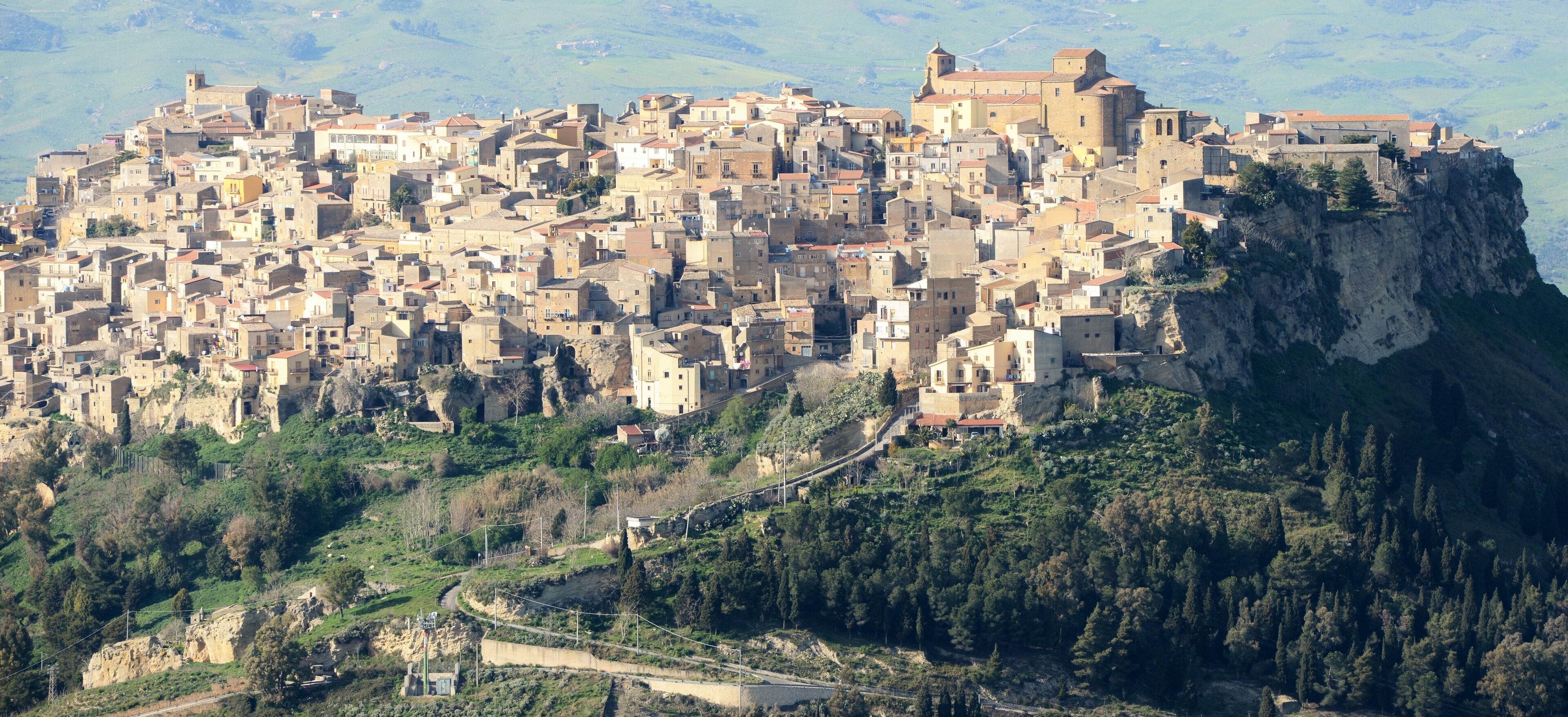 A densely packed medieval town perched on a cliff, surrounded by lush greenery and a winding road.