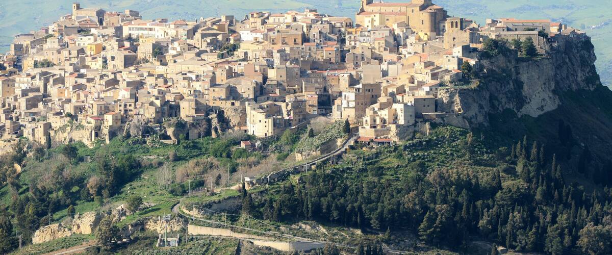 A densely packed medieval town perched on a cliff, surrounded by lush greenery and a winding road.