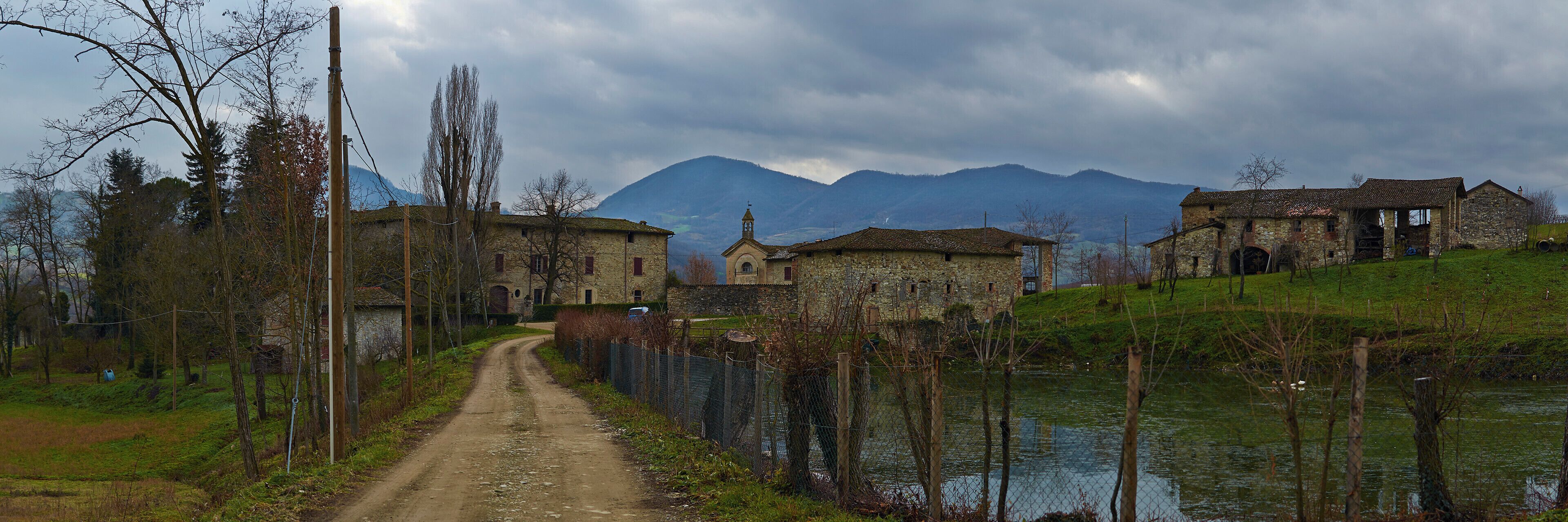A rural path leading to stone buildings, a body of water, and mountains in the background.