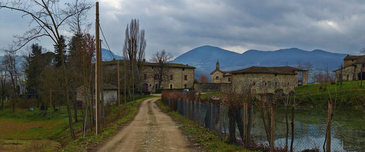 A rural path leading to stone buildings, a body of water, and mountains in the background.