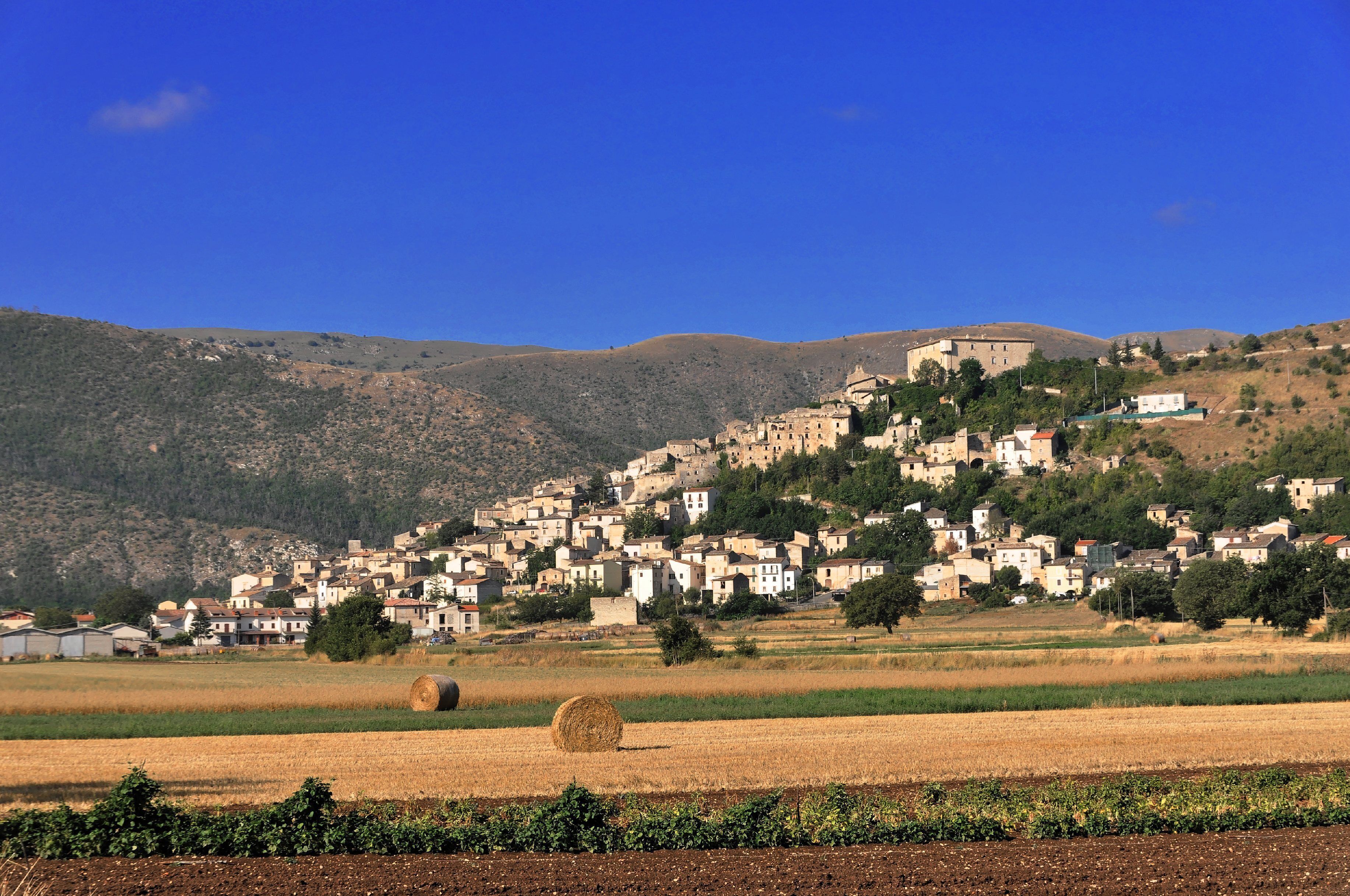 A hillside village with buildings, a large hill in the background, and a field with hay bales.