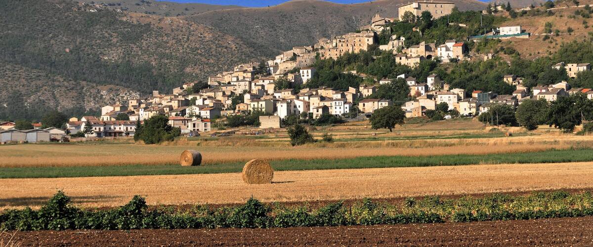 A hillside village with buildings, a large hill in the background, and a field with hay bales.