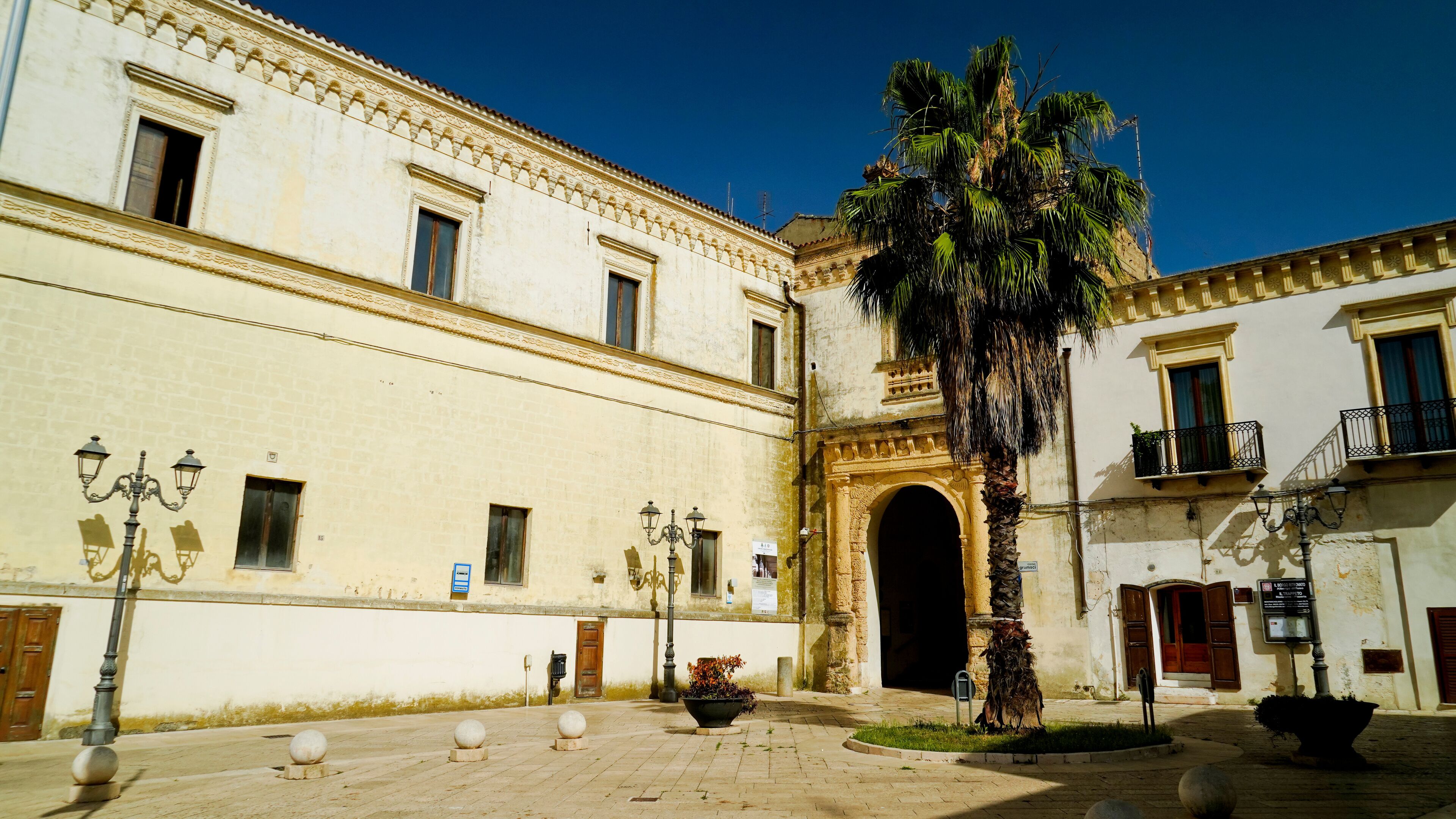 A historic building with a large archway, a palm tree, and a courtyard with bollards and a planter.