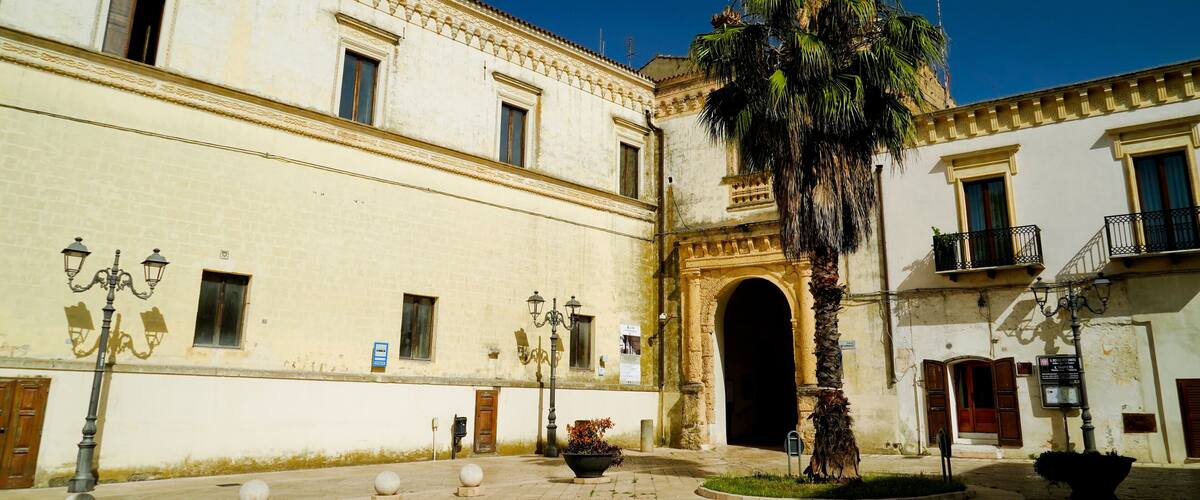 A historic building with a large archway, a palm tree, and a courtyard with bollards and a planter.