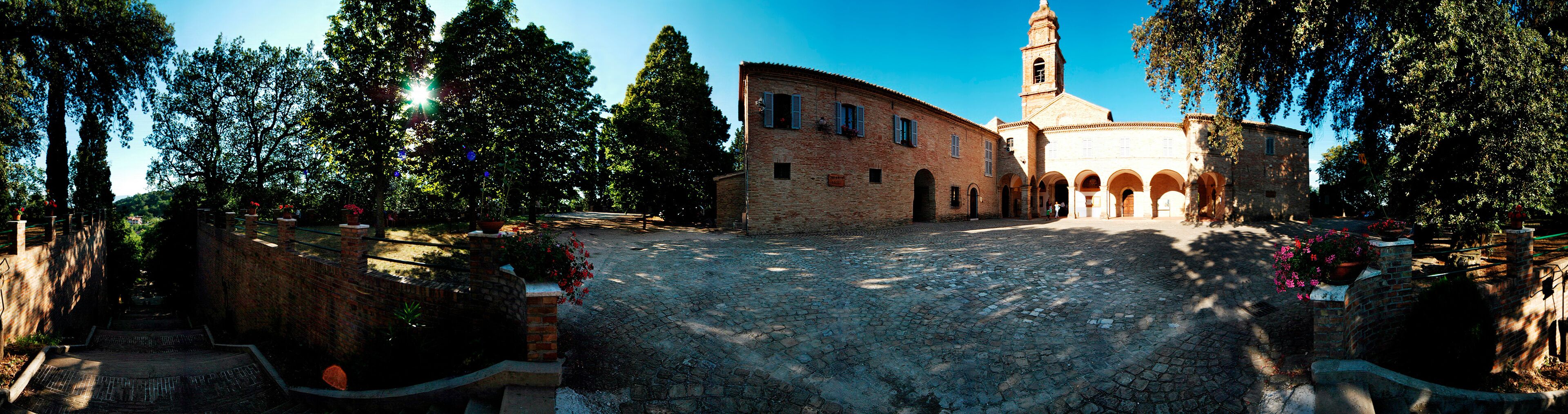 A cobblestone street leading to a historic building with a tower, surrounded by trees and flower beds.
