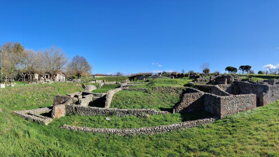 Ancient ruins on a grassy hill with stone walls and pathways, surrounded by trees and a clear blue sky.