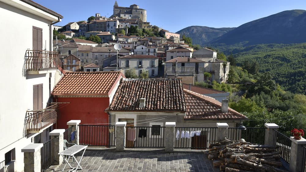 A hillside town with terracotta-roofed buildings, a prominent castle, and lush green mountains in the background.
