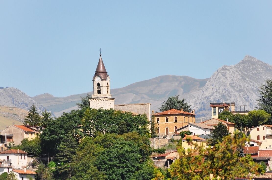 A town with a prominent church tower, surrounded by residential buildings and mountains in the background.